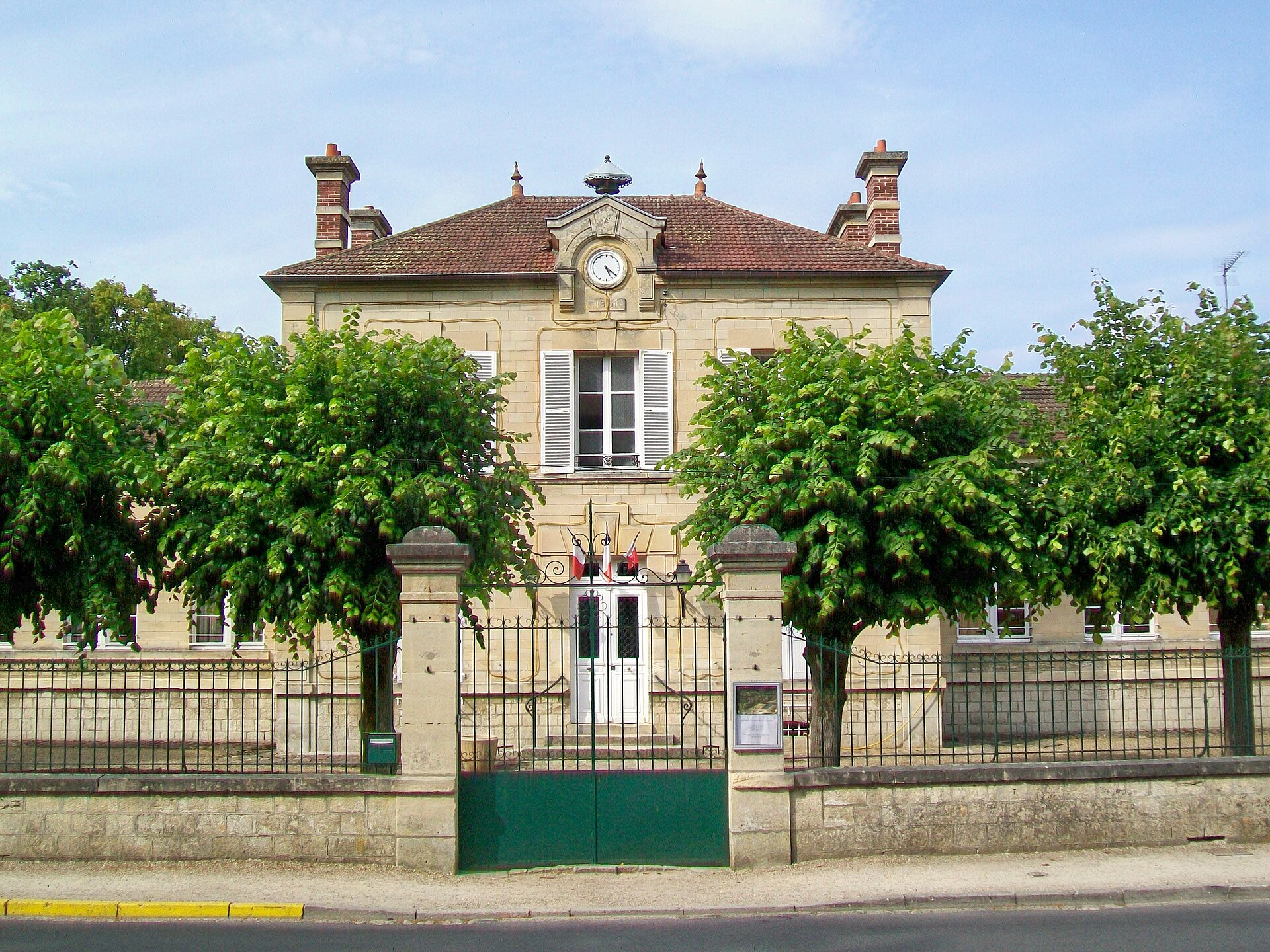 La façade de la mairie, un bâtiment de pierre à un étage derrière une grille et deux arbres